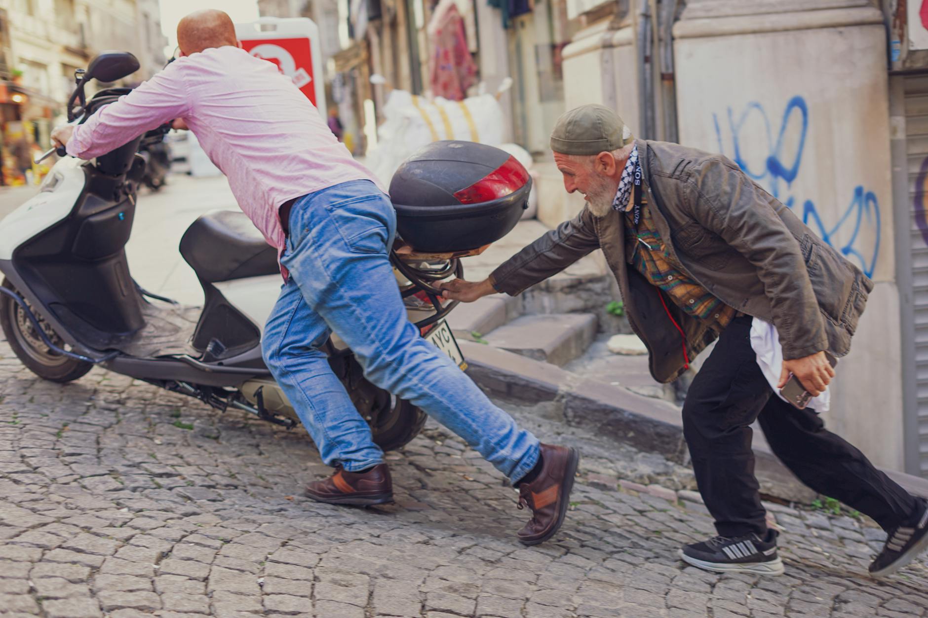 Dwóch mężczyzn pcha skuter pod górę po brukowanej ulicy