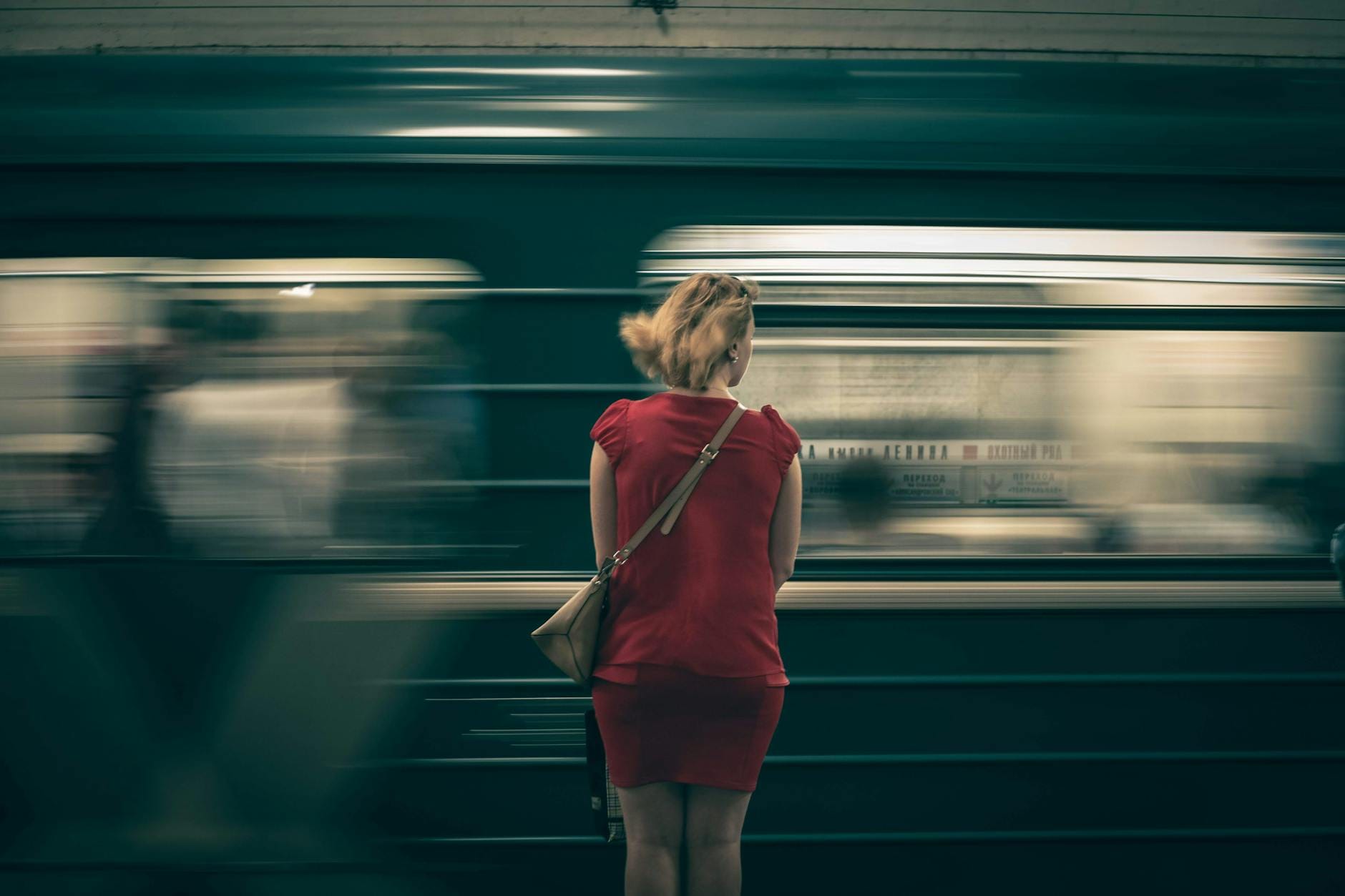 Kobieta w czerwonej sukience czeka na peronie, mijają ją rozmyte wagony metra